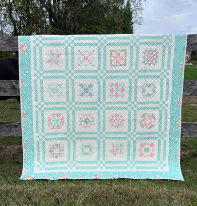 A handmade patchwork quilt with a pale blue and white checkerboard pattern and floral star motifs, hanging outdoors on a wooden fence in a grassy yard.