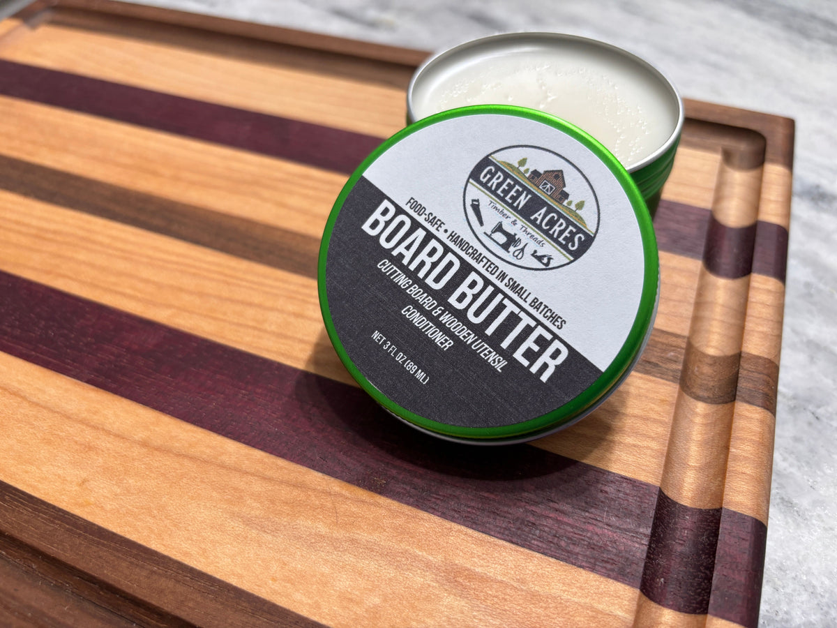 A tin of Green Acres Board Butter conditioner resting on a striped wooden cutting board next to an open tin showing the white balm inside.