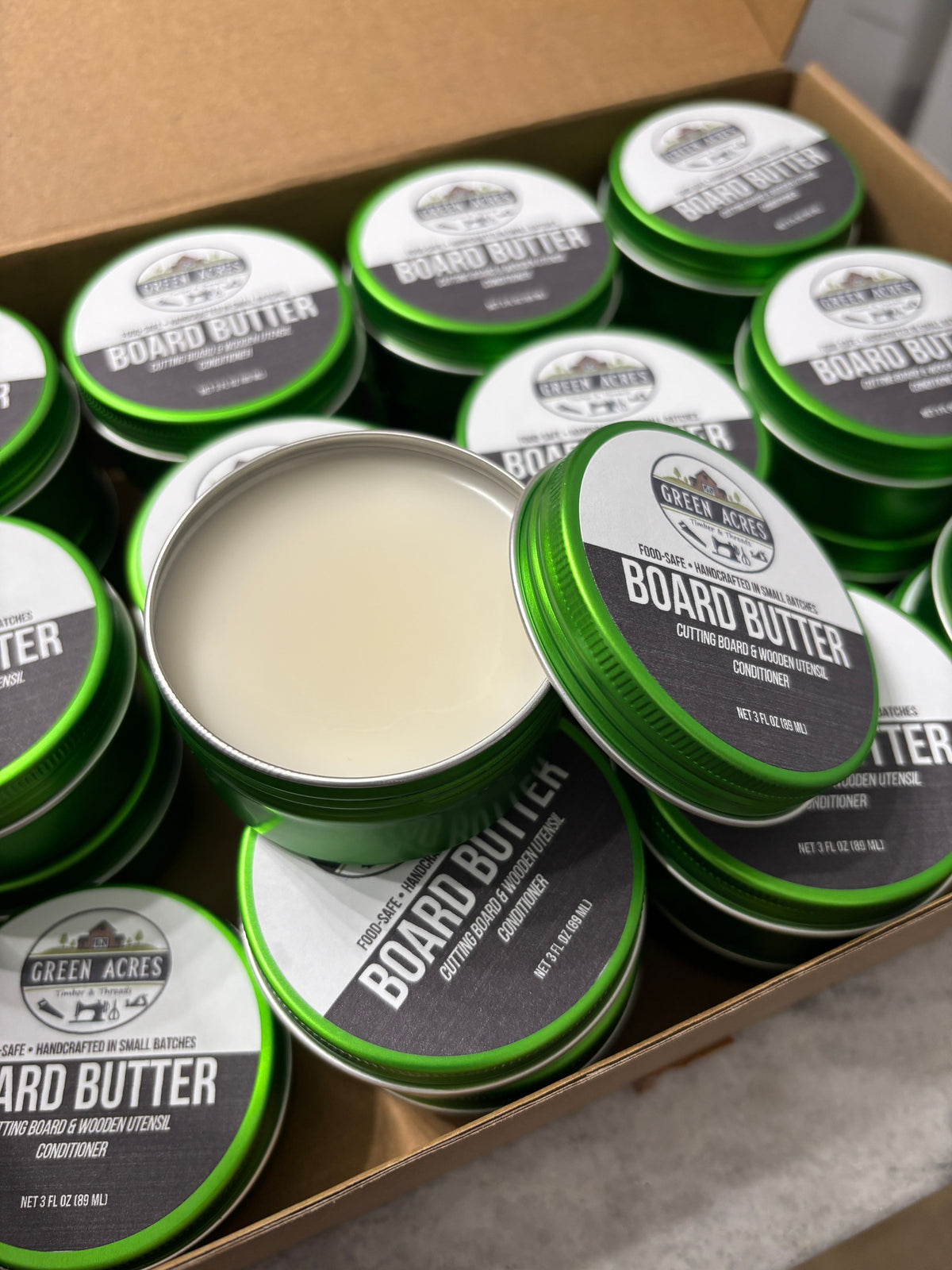 A box of Green Acres Board Butter wood conditioner, showing multiple green metal tins, with one open tin displaying the off-white conditioner cream.