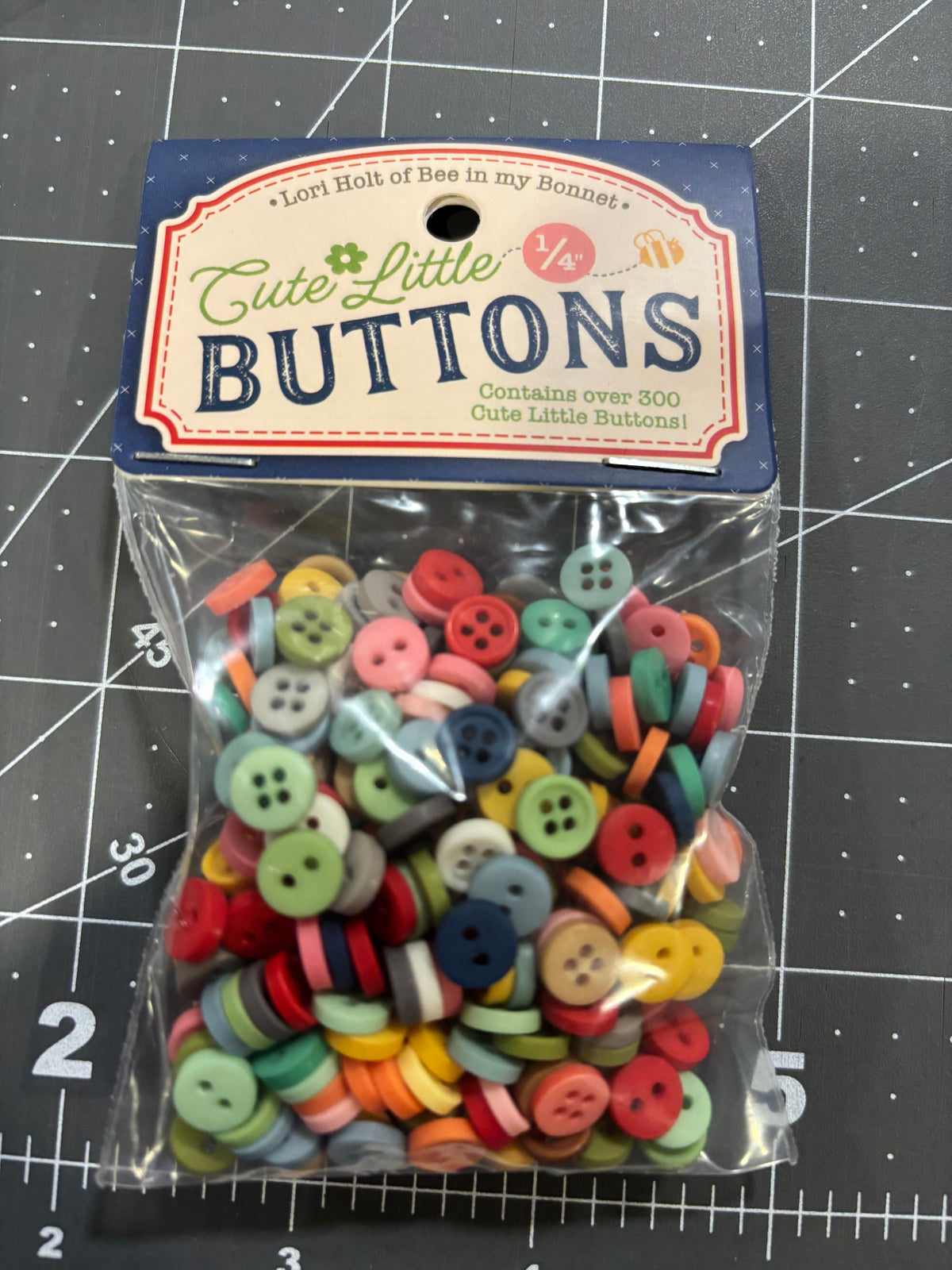 A clear plastic bag filled with many small decorative buttons by Lori Holt in assorted colors, attached to a blue and cream paper header card that reads 'Cute-Little BUTTONS.' The package is photographed on a dark gray gridded cutting mat background.