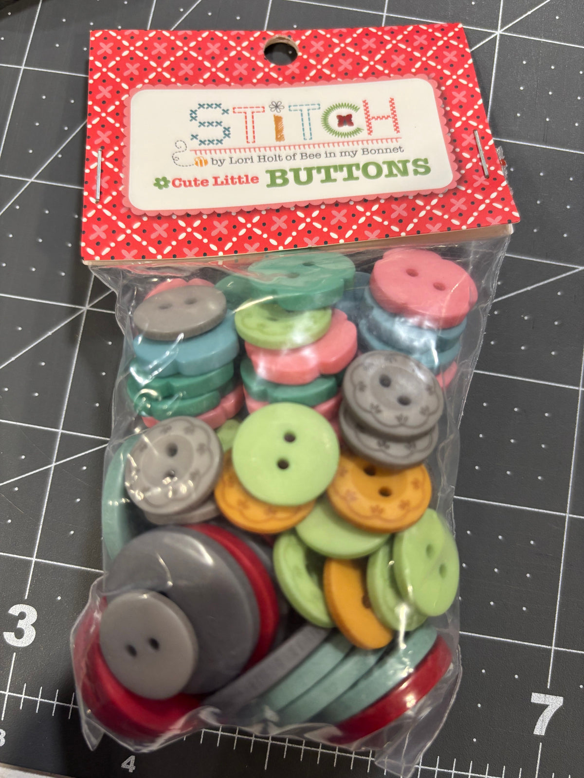 A clear plastic bag filled with assorted round craft buttons in multiple colors, sealed with a red patterned header card labeled 'STITCH Cute Little BUTTONS' by Lori Holt of Bee in my Bonnet, placed on a gray cutting mat.