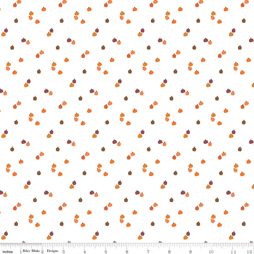 White fabric with a repeating pattern of small orange pumpkins and brown acorns, shown flat with a ruler along the bottom edge for scale.