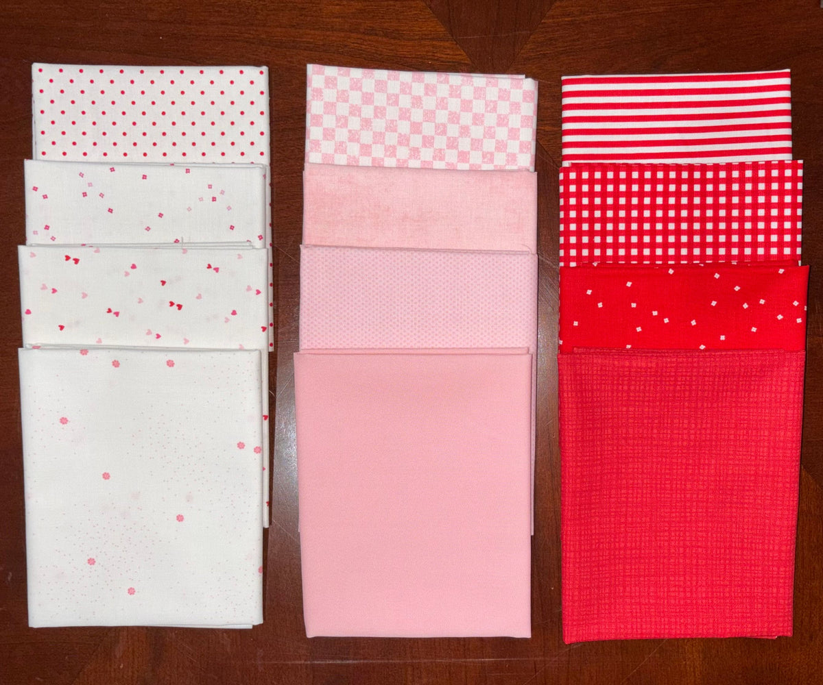 Three stacks of folded fabric samples in pink, red, and white patterns, arranged side by side on a dark wood background.