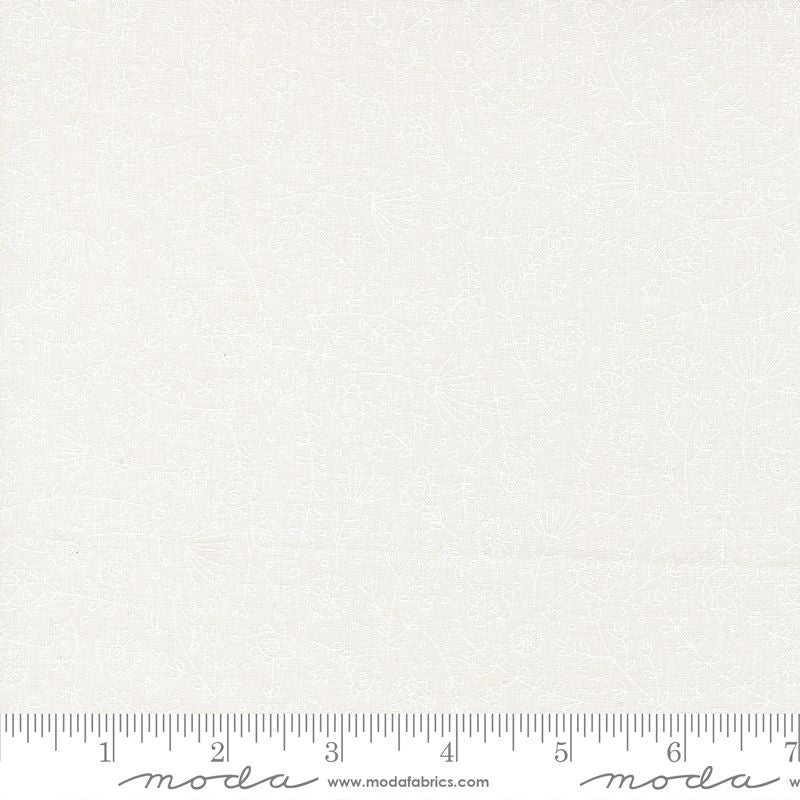 A close-up of a white-on-white fabric with a subtle textured floral pattern, featuring a ruler at the bottom edge for scale with Moda branding.