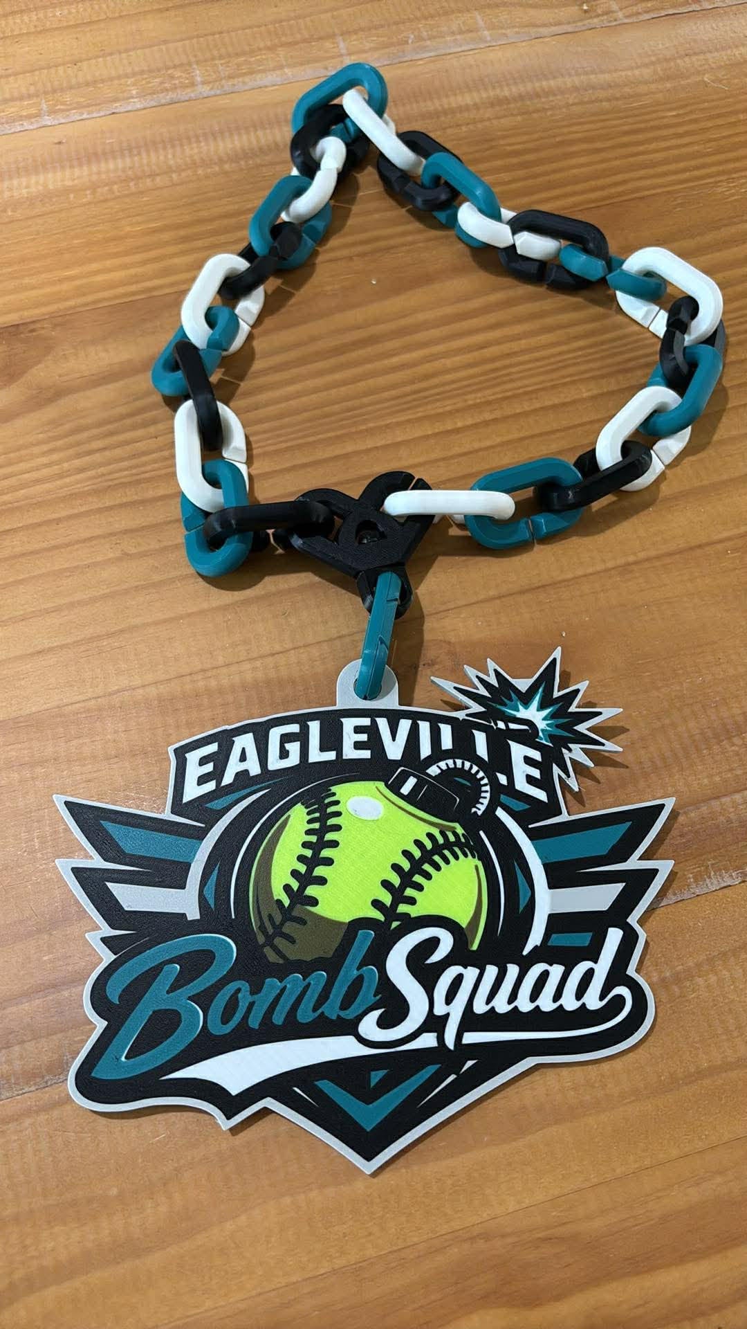 A black, teal, and white chain accessory with an 'Eagleville Bomb Squad' softball graphic charm, displayed on a wooden surface.