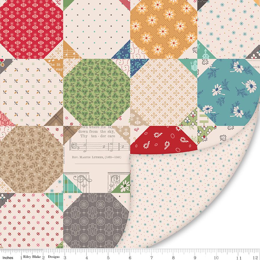 A close-up of a quilted fabric design showing multiple patterned square and hexagonal blocks in red, green, blue, beige, and white, with a ruler along the bottom edge showing measurements.