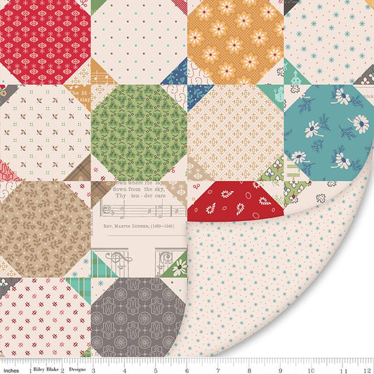 A close-up of a quilted fabric design showing multiple patterned square and hexagonal blocks in red, green, blue, beige, and white, with a ruler along the bottom edge showing measurements.