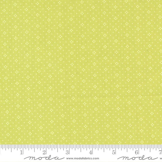 A close-up of a fabric pattern featuring a light yellow-green background with a repeating small white floral or starburst motif. A white ruler along the bottom edge shows measurement markings and the Moda Fabrics brand name.