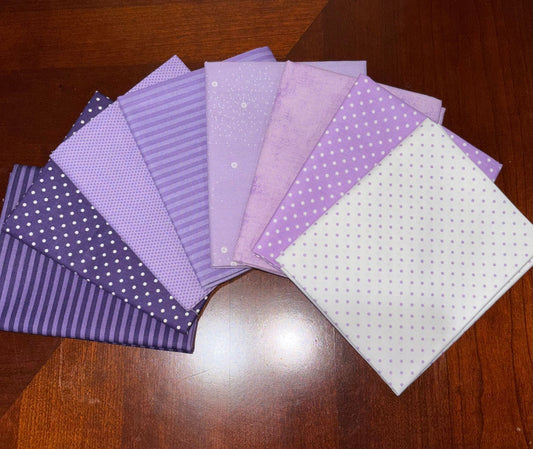 A fan arrangement of purple patterned fabric squares, including solid lavender, white polka dots, and dark purple stripes, displayed on a dark wood tabletop.