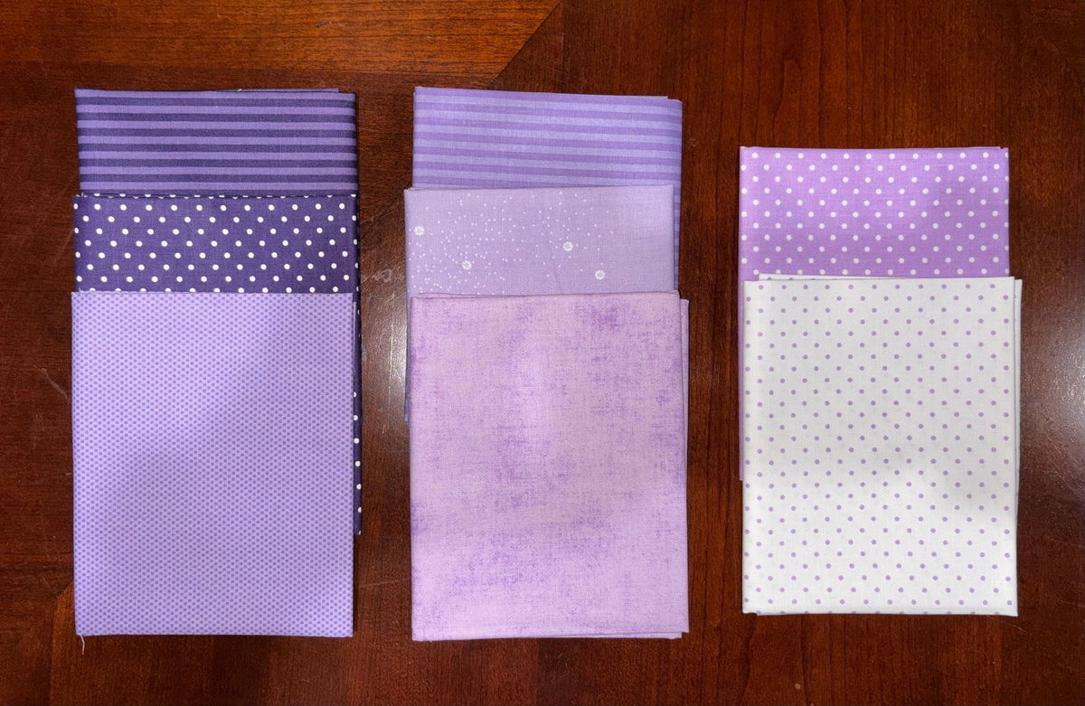 Three stacks of purple and lavender fabric swatches with different patterns (solid, polka dot, striped) arranged side-by-side on a dark wooden table.
