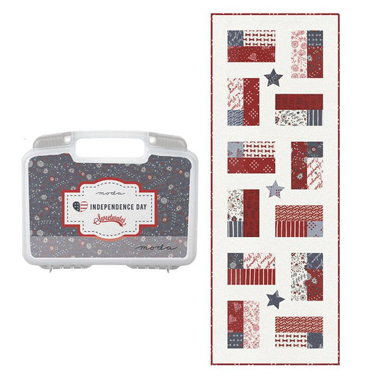 Quilt kit with a plastic reusable case labeled 'Independence Day' on a white background next to a quilted patriotic table runner with red, white and blue fabric.