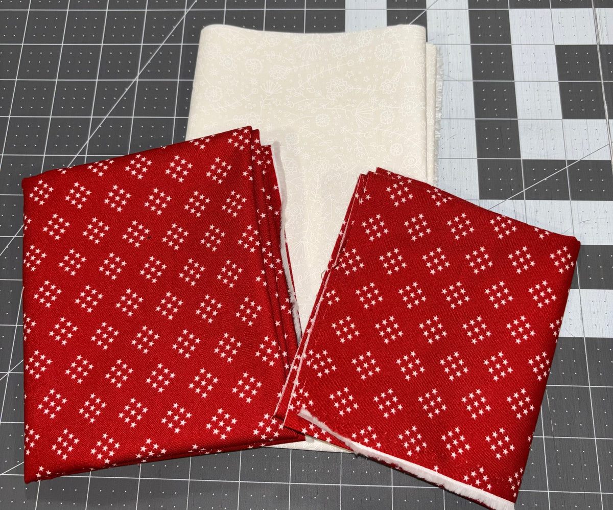 Two folded pieces of red fabric with a white geometric star pattern are displayed next to a folded piece of off-white fabric on a gray cutting mat.