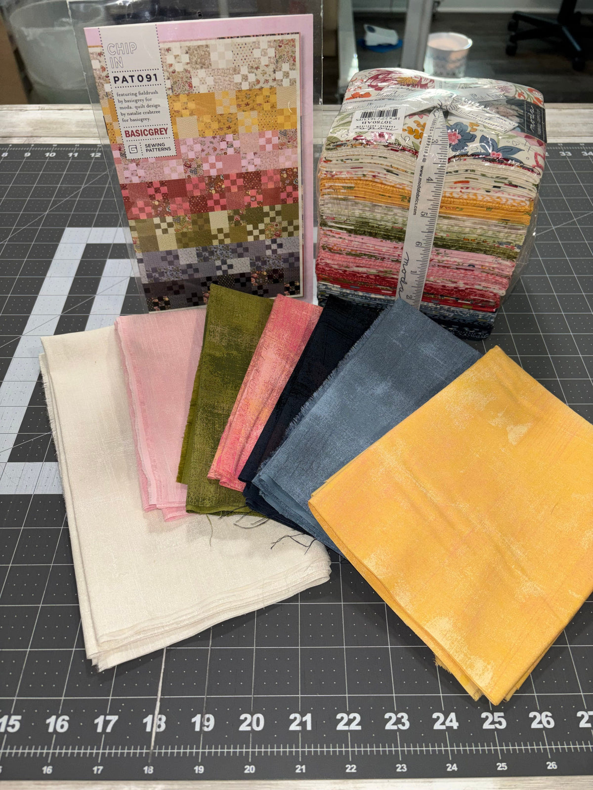A fat quarter bundle and some folded stacks of grunge fabric displayed on a black gridded cutting mat. The fabrics include various colors and patterns, such as solid yellow, pink, green, navy, and a multicolor pixelated print.