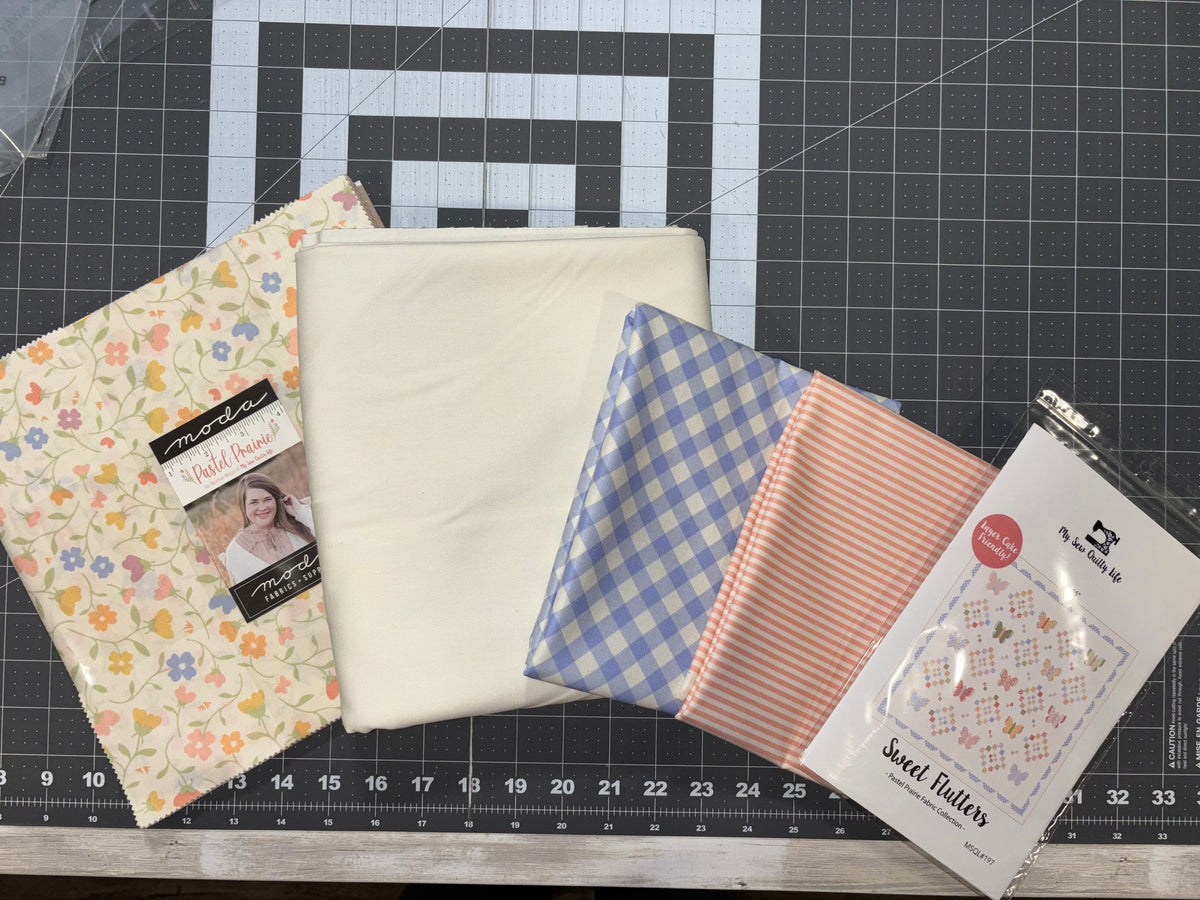 A layer cake and three sewing fabric bundles and a packaged quilt pattern arranged on a black gridded cutting mat.