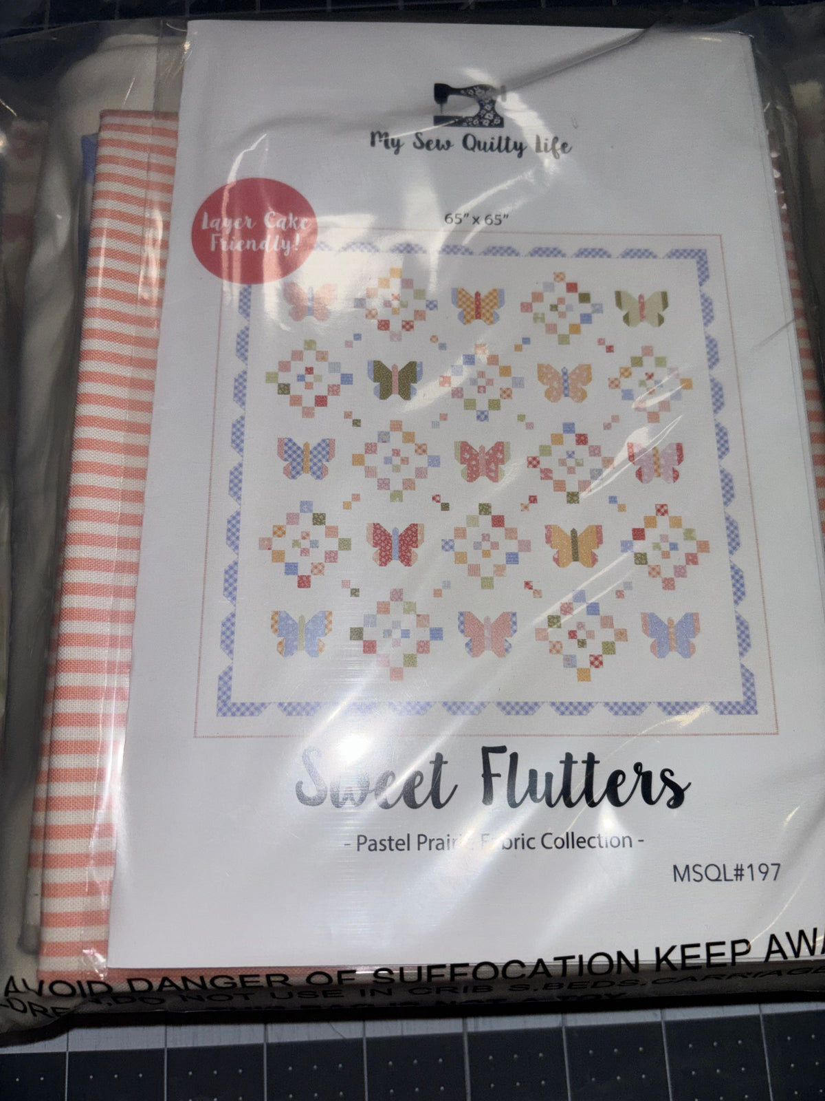 A packaged quilt kit titled 'Sweet Flutters' from the 'Pastel Prairie Collection,' showing a butterfly quilt design on the cover, photographed inside clear plastic wrapping.