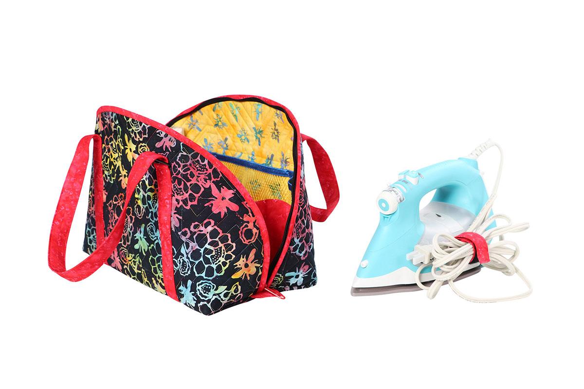 A fabric bag with a black background and colorful floral and bee patterns, shown open with red handles, alongside a light blue iron on a white background.