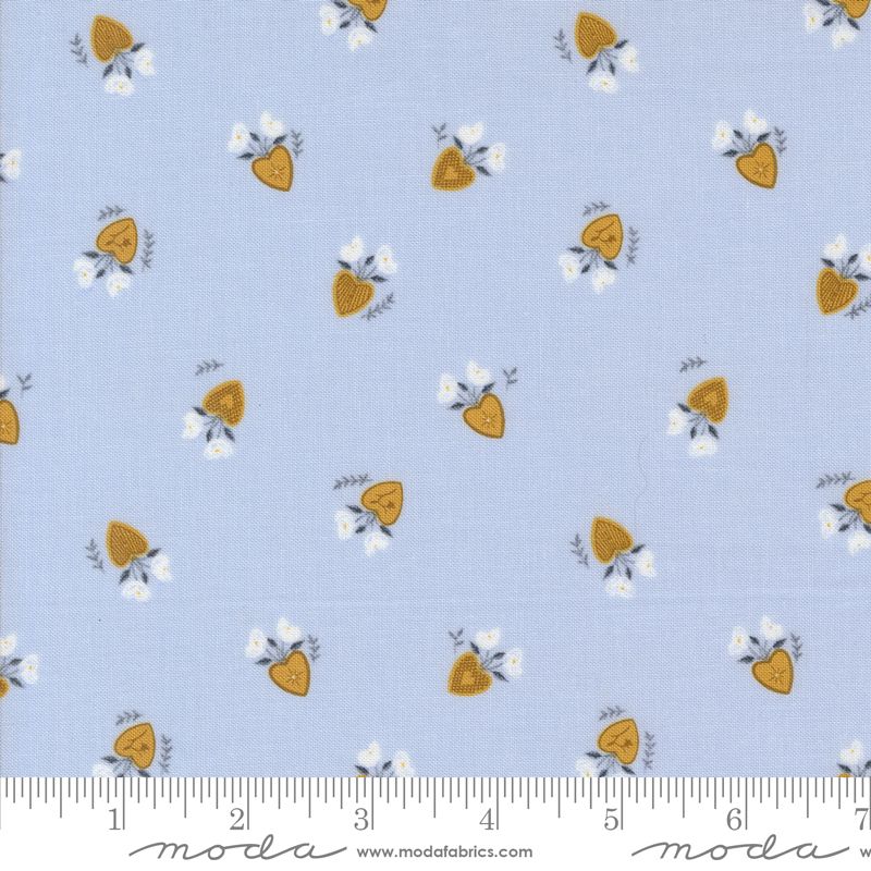 Light blue fabric with a repeating pattern of small yellow-orange hearts, white flowers, and gray leaves, shown with a measuring ruler along the bottom edge. The brand Modafabrics is printed on the ruler.