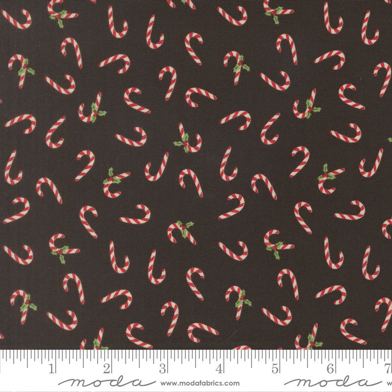 A close-up of a fabric pattern featuring red and white striped candy canes with small green holly leaves on a dark brown background, with a white ruler along the bottom edge showing scale.
