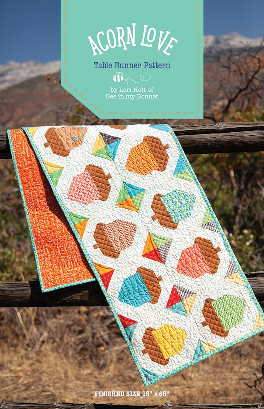 A quilted table runner pattern with an autumn acorn table runner shown draped over a wooden fence outdoors, with a teal banner at the top reading 'Acorn Love Table Runner Pattern' and a mountain landscape in the background.