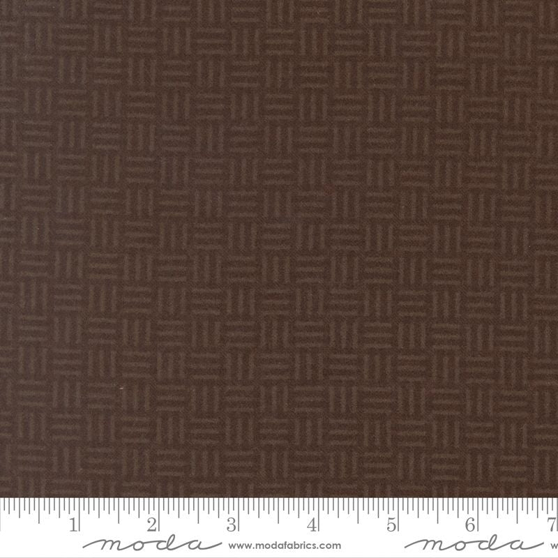 A close-up of dark brown fabric with a repeating geometric basketweave pattern, with a white ruler along the bottom edge showing measurements. The brand name 'Moda Fabrics' is visible on the ruler.