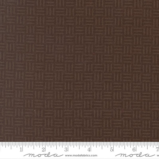 A close-up of dark brown fabric with a repeating geometric basketweave pattern, with a white ruler along the bottom edge showing measurements. The brand name 'Moda Fabrics' is visible on the ruler.