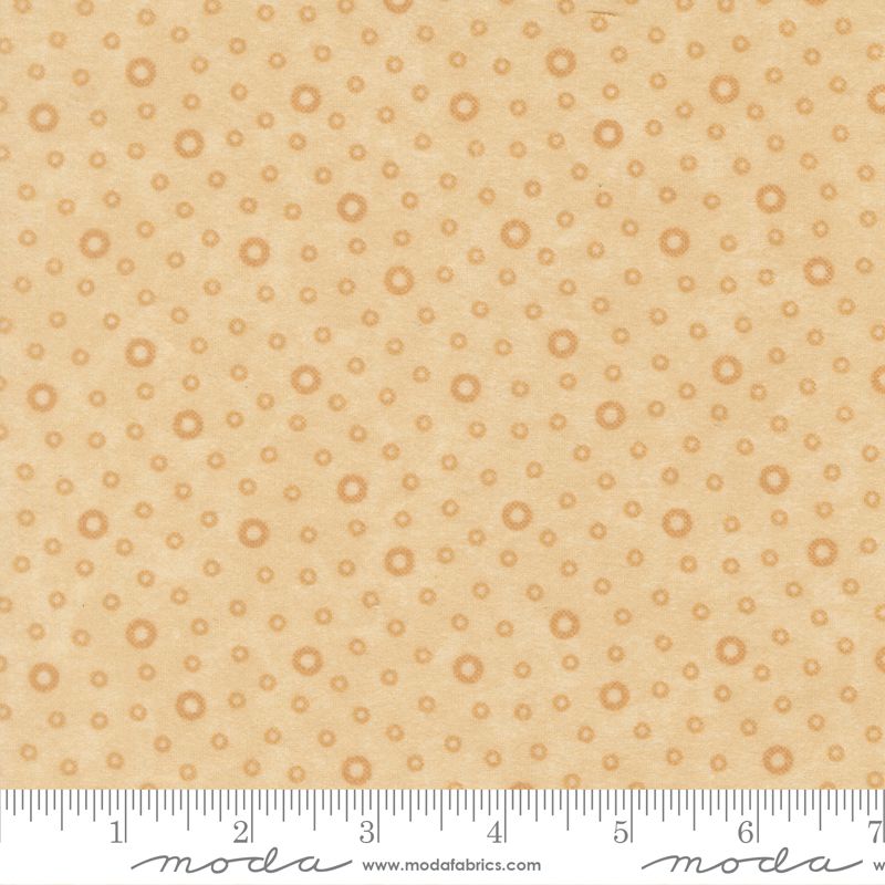A close-up of a fabric pattern featuring small tan circular motifs on a warm beige background, with a ruler along the bottom edge showing scale and Modas Fabrics branding.