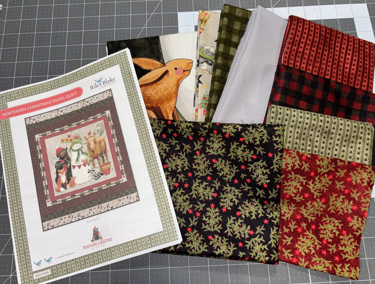 A quilt pattern booklet titled 'Northern Christmas Village Quilt' by Riley Blake Designs is displayed alongside multiple fabric swatches showcasing the various patterns included in the quilt design, arranged on a gray gridded cutting mat.