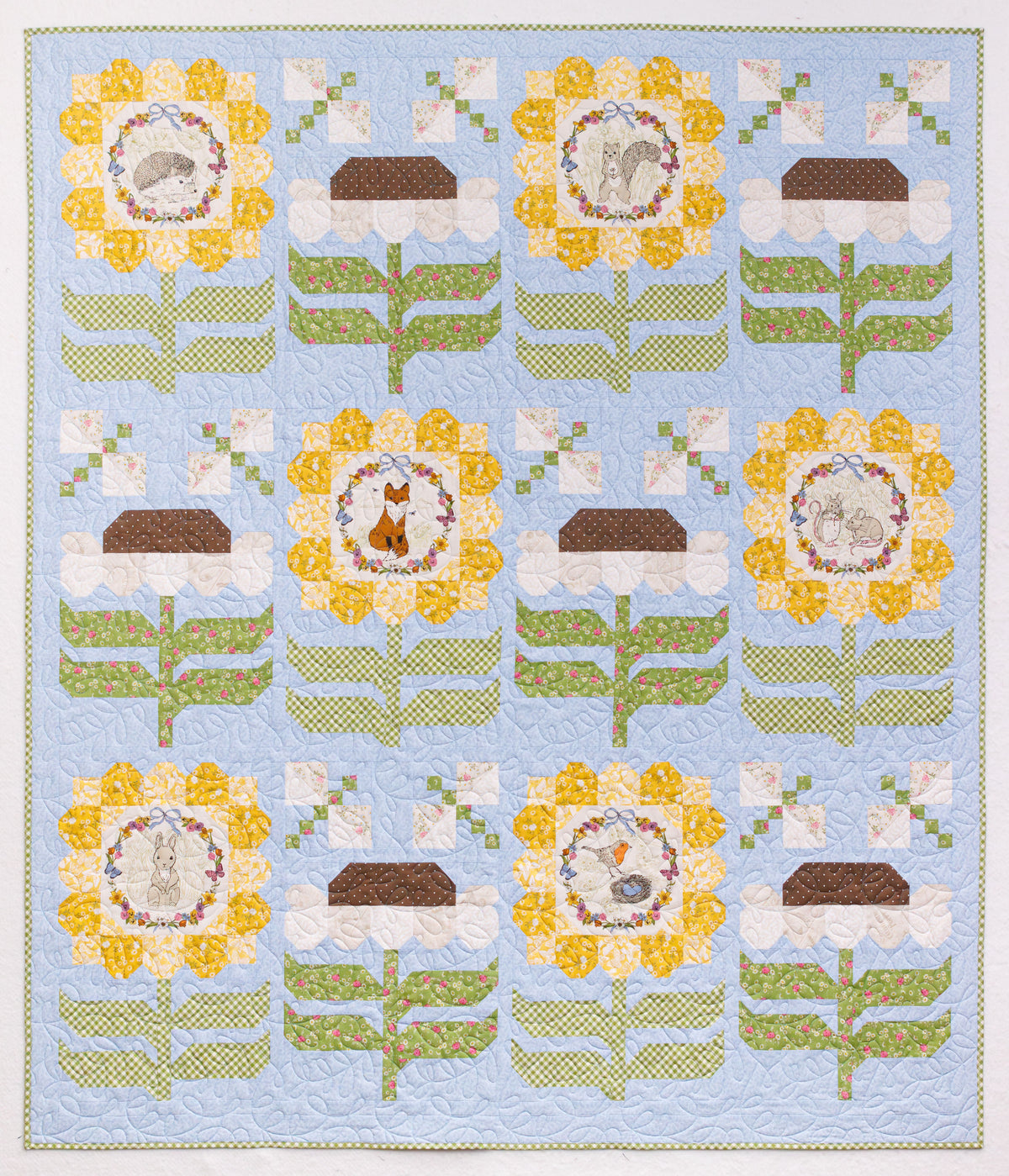 A square quilt featuring a repeating floral and animal motif on a light blue background. The design includes yellow flowers with white centers, white blossoms with green stems, and circular motifs containing small animals, a hedgehog, squirrel, fox, mice, rabbit, and birds.