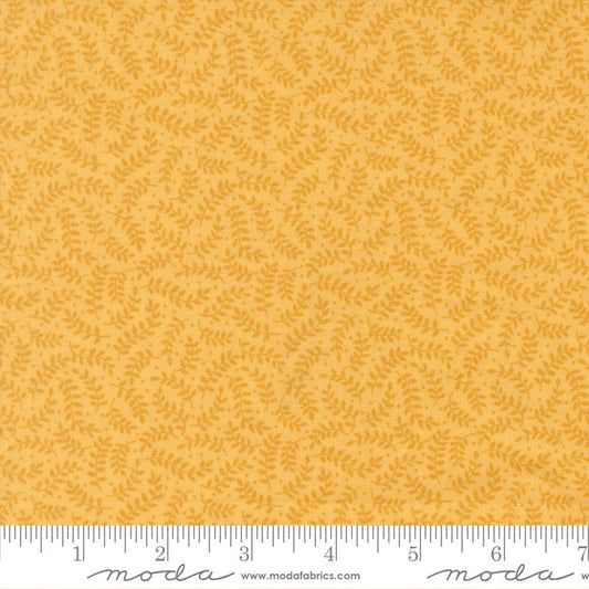 A close-up of a fabric pattern showing a warm mustard yellow background with a repeating small leafy vine motif in a slightly darker golden yellow. A white ruler with black markings is visible along the bottom edge, and the Moda Fabrics logo and website are printed at the bottom.