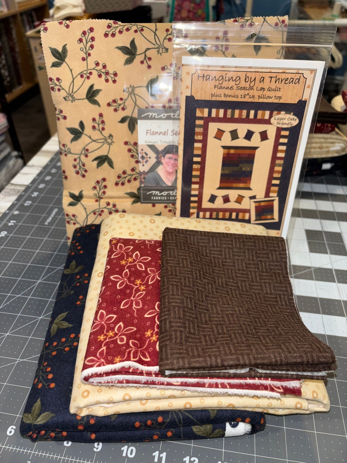 A quilting kit fabric bundle displayed on a cutting mat, including patterned quilts, folded fabrics in navy, red, beige, and brown, and a printed pattern booklet titled 'Handling by a Thread'.