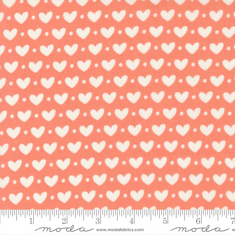 A close-up of a fabric pattern featuring repeating white hearts and small white dots on a coral pink background, with a ruler along the bottom edge showing scale and a Moda Fabrics watermark.