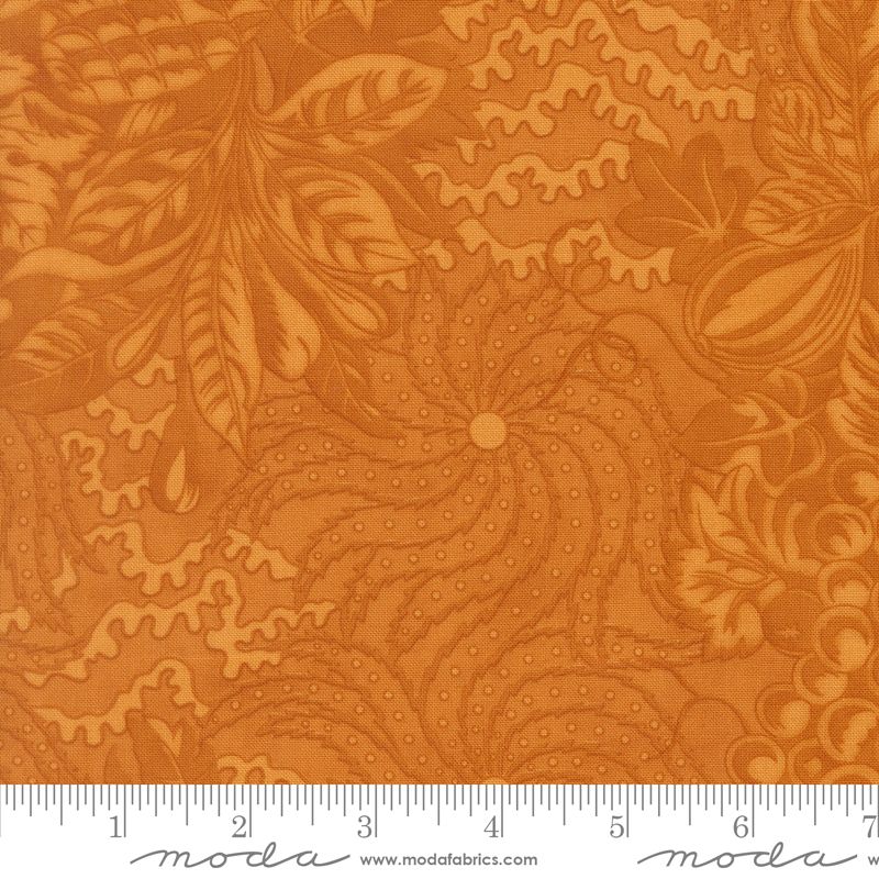 A close-up of butterscotch fabric with a floral and leaf pattern, shown with a measuring ruler along the bottom edge. The brand name 'Moda' is visible on the ruler.