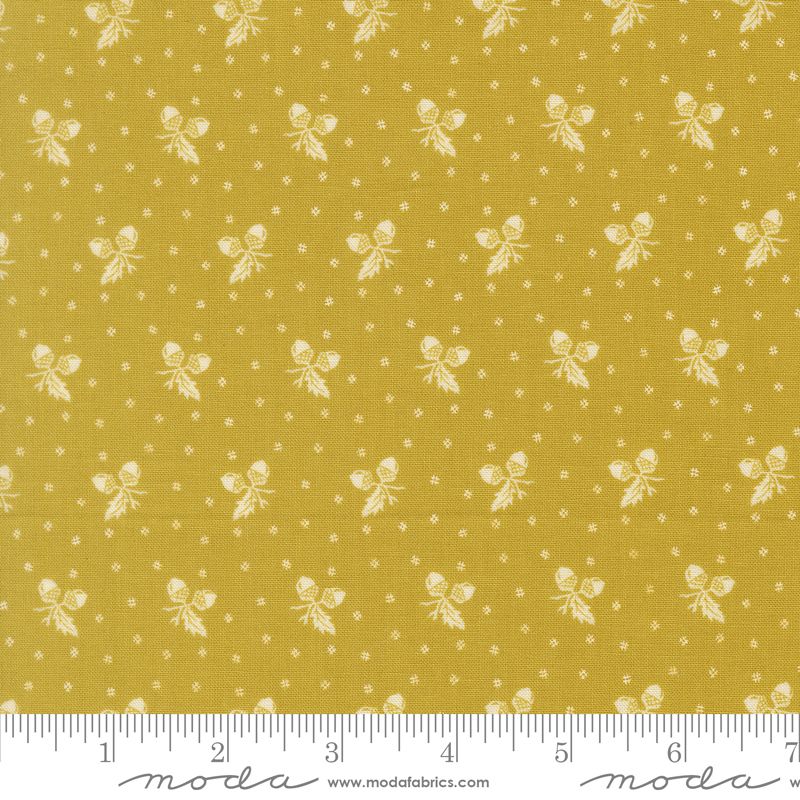 A close-up of a mustard yellow fabric with a repeating white pattern of small bows, leaves, and tiny dots, with a white ruler along the bottom edge showing measurements. The brand name 'modafabrics' is printed on the ruler.
