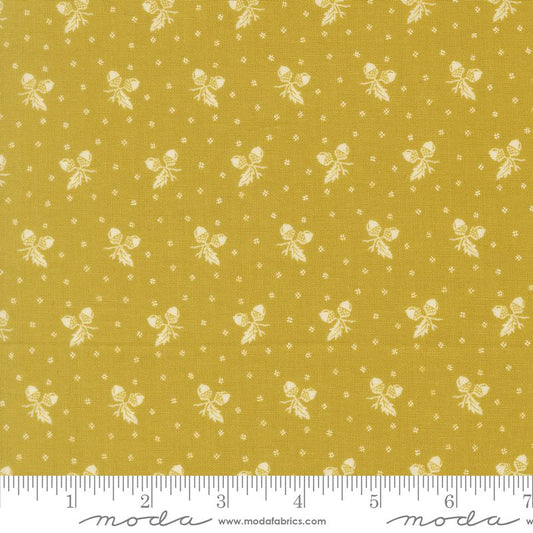 A close-up of a mustard yellow fabric with a repeating white pattern of small bows, leaves, and tiny dots, with a white ruler along the bottom edge showing measurements. The brand name 'modafabrics' is printed on the ruler.