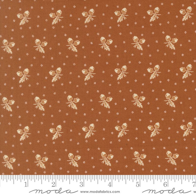 A close-up of a fabric pattern featuring a warm rust-orange background with a repeating cream acorn and leaf motif, small white dotted diamond motifs, and a white ruler along the bottom edge showing measurements. The brand name 'moda' is visible on the ruler watermark.