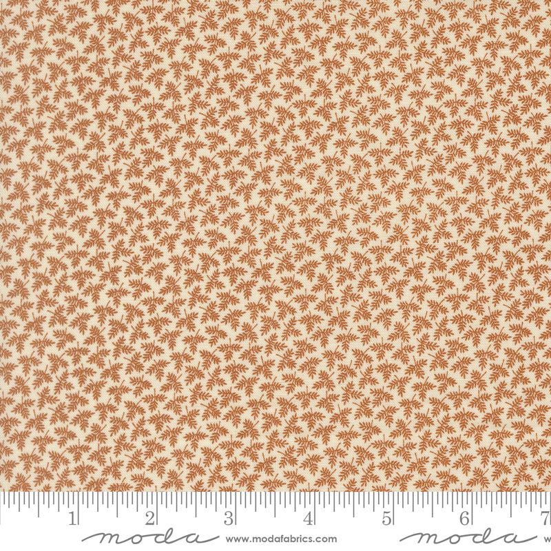 A close-up of a fabric pattern showing a repeating small brown palm-leaf or fan-like motif on a warm cream background, with a ruler along the bottom edge for scale. Moda fabric.
