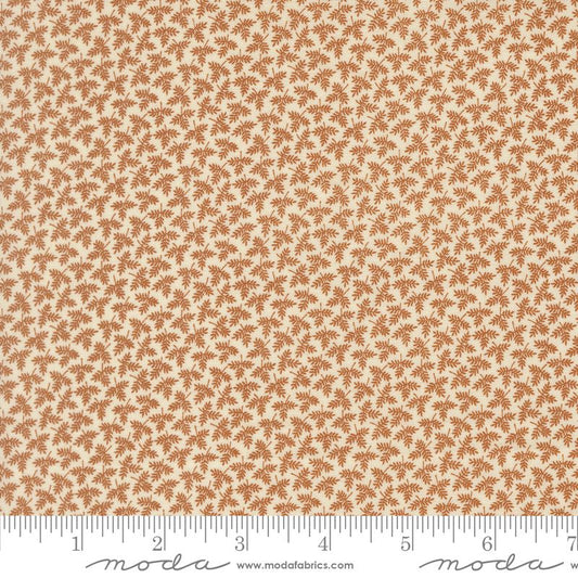 A close-up of a fabric pattern showing a repeating small brown palm-leaf or fan-like motif on a warm cream background, with a ruler along the bottom edge for scale. Moda fabric.

