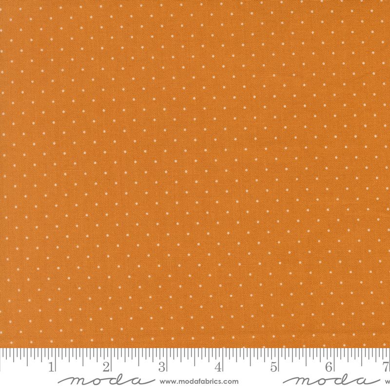 A close-up fabric swatch showing an orange background with a repeating pattern of small white dots. A white ruler along the bottom edge shows measurement markings, and the Moda Fabrics logo and website appear at the bottom.