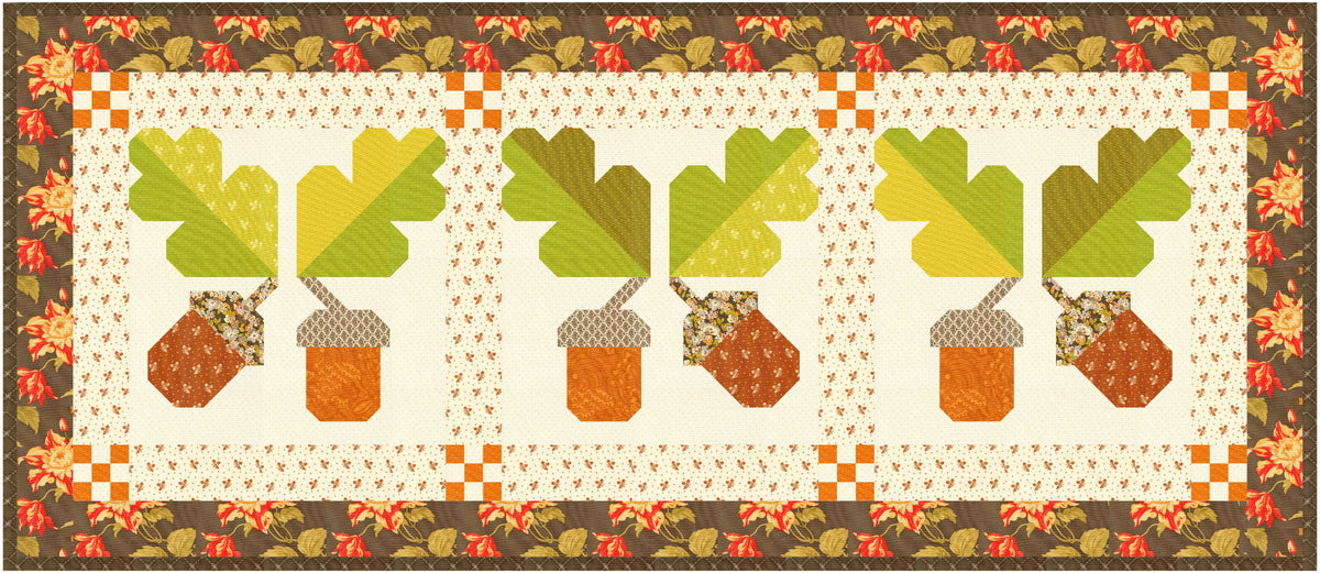 A quilted table runner featuring three repeating acorn and leaf motifs in autumn colors, framed by a floral border. The design includes green oak leaves, brown acorns, and a cream center with a patterned border.