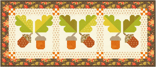 A quilted table runner featuring three repeating acorn and leaf motifs in autumn colors, framed by a floral border. The design includes green oak leaves, brown acorns, and a cream center with a patterned border.