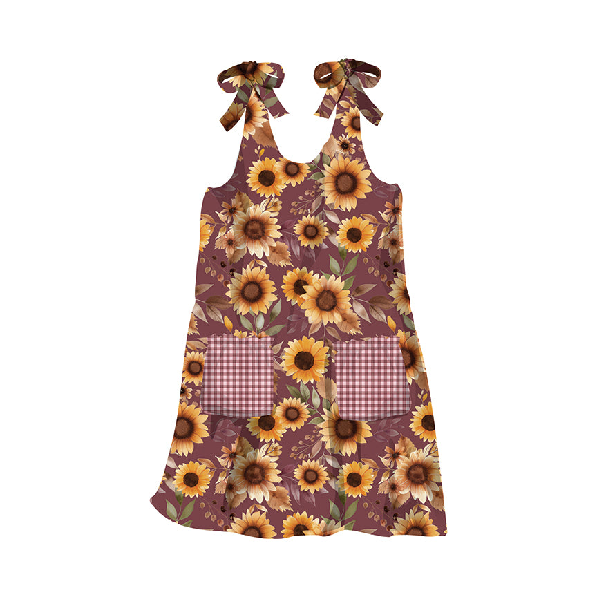 A sleeveless dress-style apron with a brown and yellow sunflower print, featuring two wine-and-white gingham pockets and matching ribbon neck ties, shown on a plain white background.
